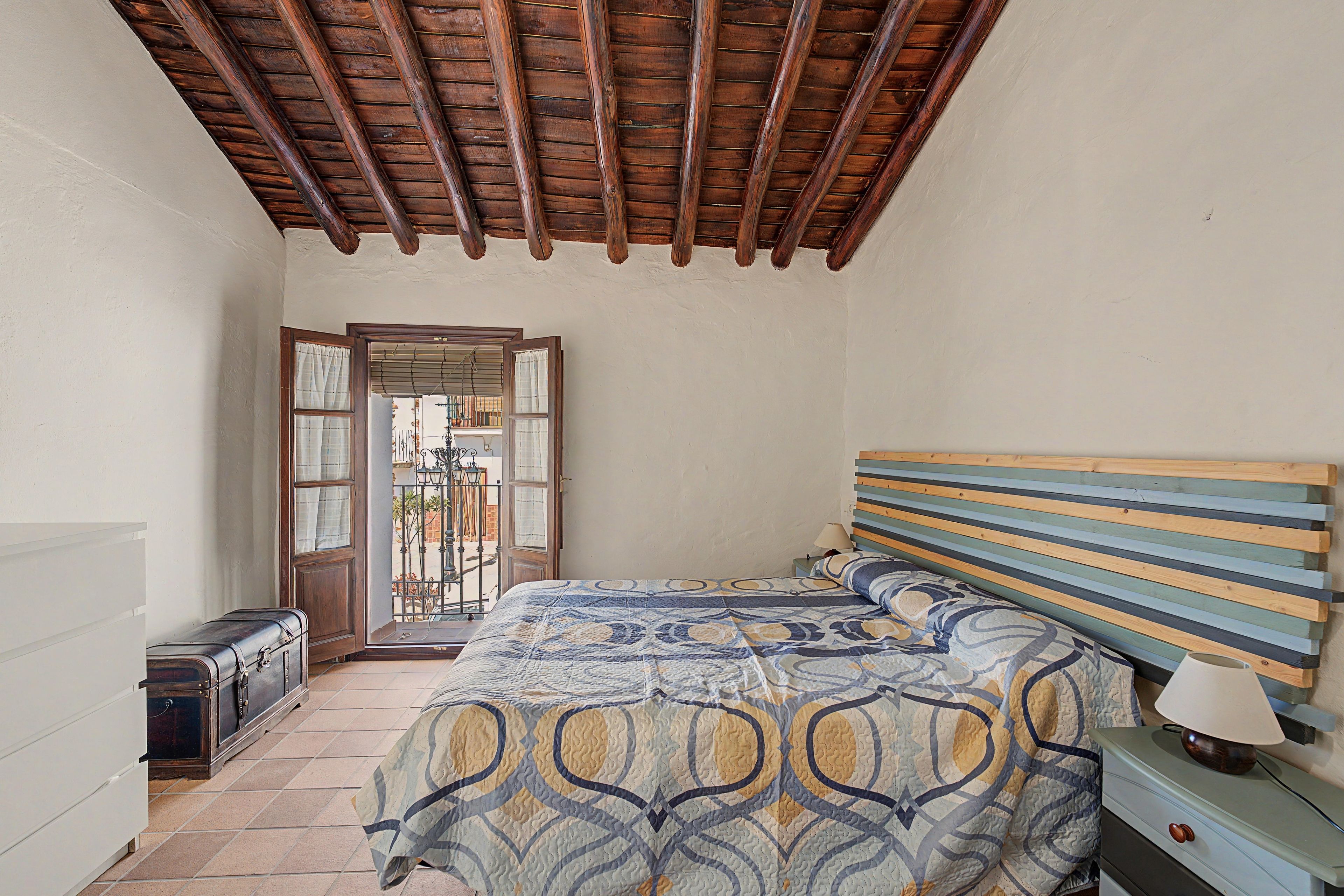 Une chambre avec un plafond en bois, un lit recouvert d’une couvre-lit à motifs, une table de chevet avec une lampe et une valise posée sur le sol.