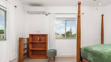 A bedroom with a four-poster bed, a wooden shelf unit, a small stool, and a window with blinds.