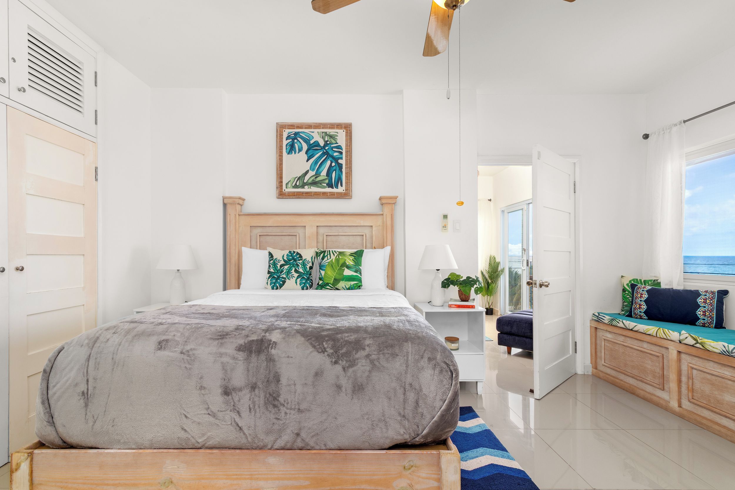 A hotel room with a large bed, a wooden headboard, a painting of tropical leaves, a nightstand with a lamp, a small table with a plant, and a view of the sea through a window.