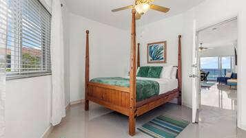 Una cama con dosel de madera y una colcha verde, un ventilador de techo y vista al océano a través de una puerta.