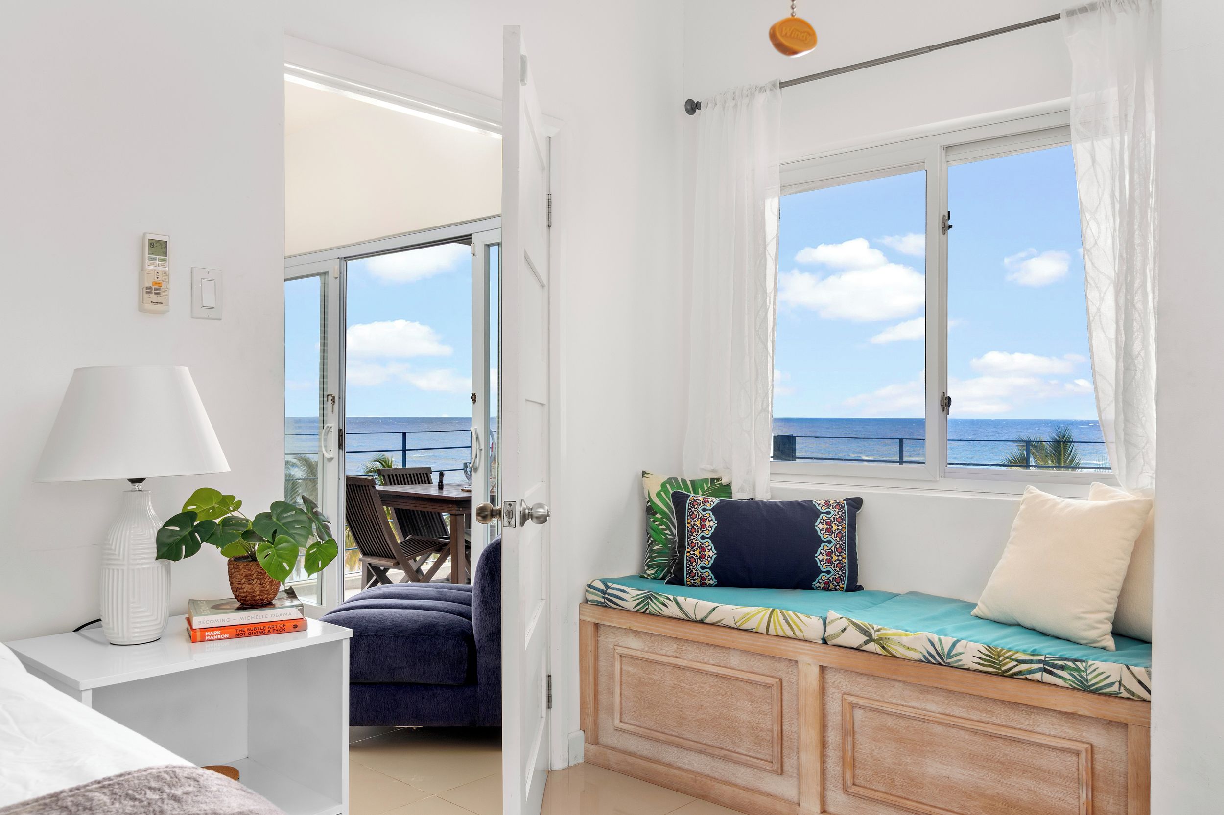 A room with a bed, a bedside table with a lamp, a window with a view of the sea, and a seating area with cushions.