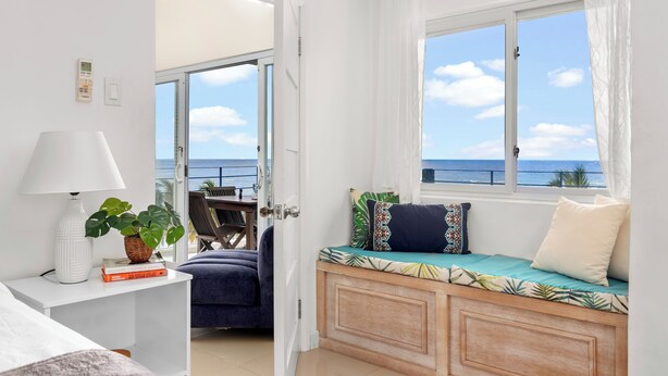 Quarto com cama, mesa de cabeceira com abajur, janela com vista para o mar e área de estar com almofadas.