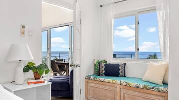 A room with a bed, a bedside table with a lamp, a window with a view of the sea, and a seating area with cushions.