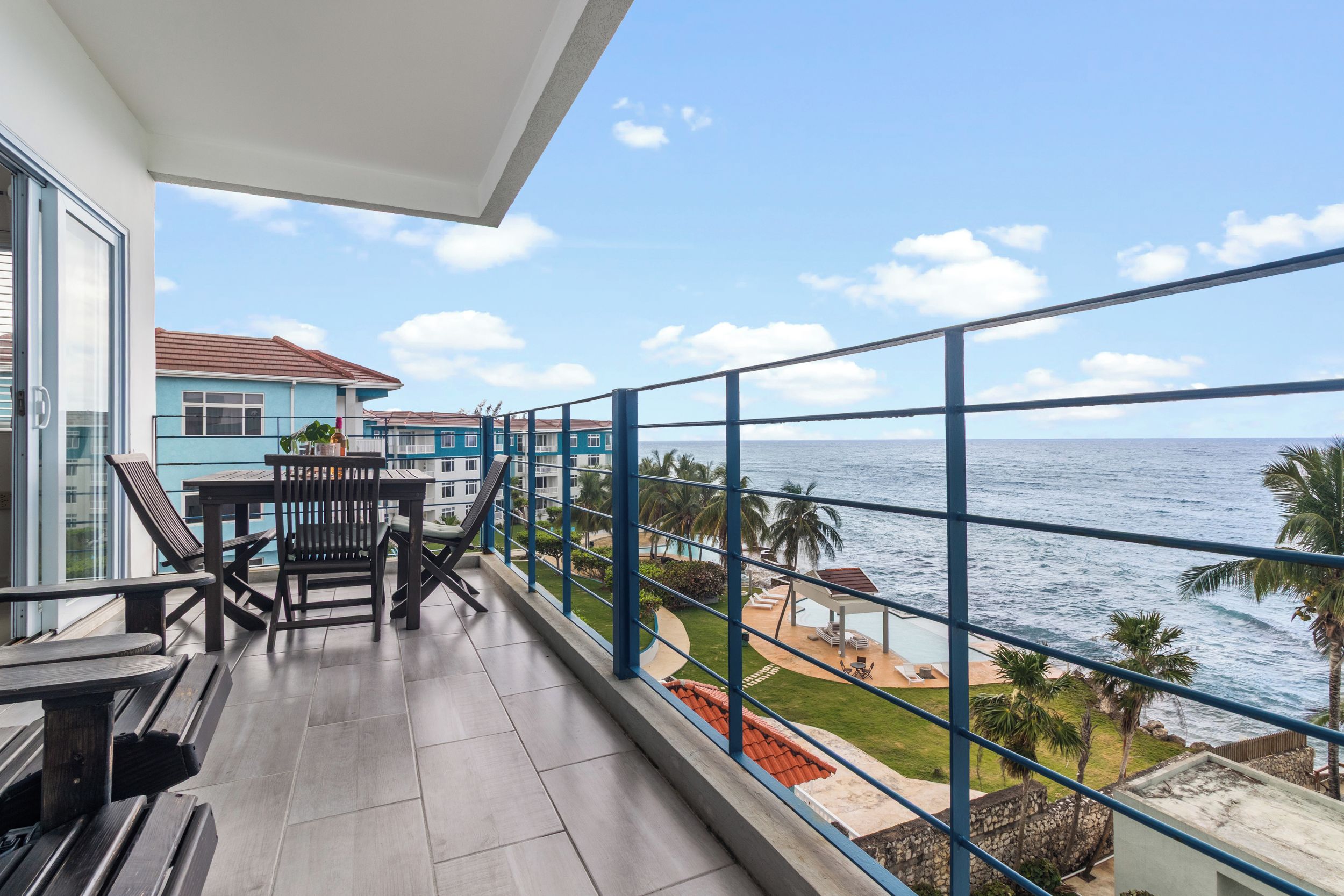 A balcony with a view of the sea, a dining table with chairs, and a building with a blue facade.