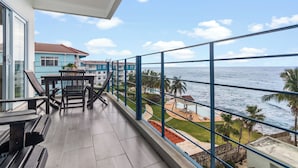 Uma varanda com vista para o mar, uma mesa de jantar com cadeiras e um edifício com fachada azul.