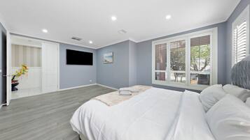 Un dormitorio con cama, televisor montado en la pared, ventana con persianas y puerta que da a otra habitación.