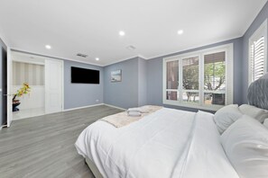 Ein Schlafzimmer mit einem Bett, einem an der Wand montierten Fernseher, einem Fenster mit Jalousien und einer Tür zu einem anderen Raum.