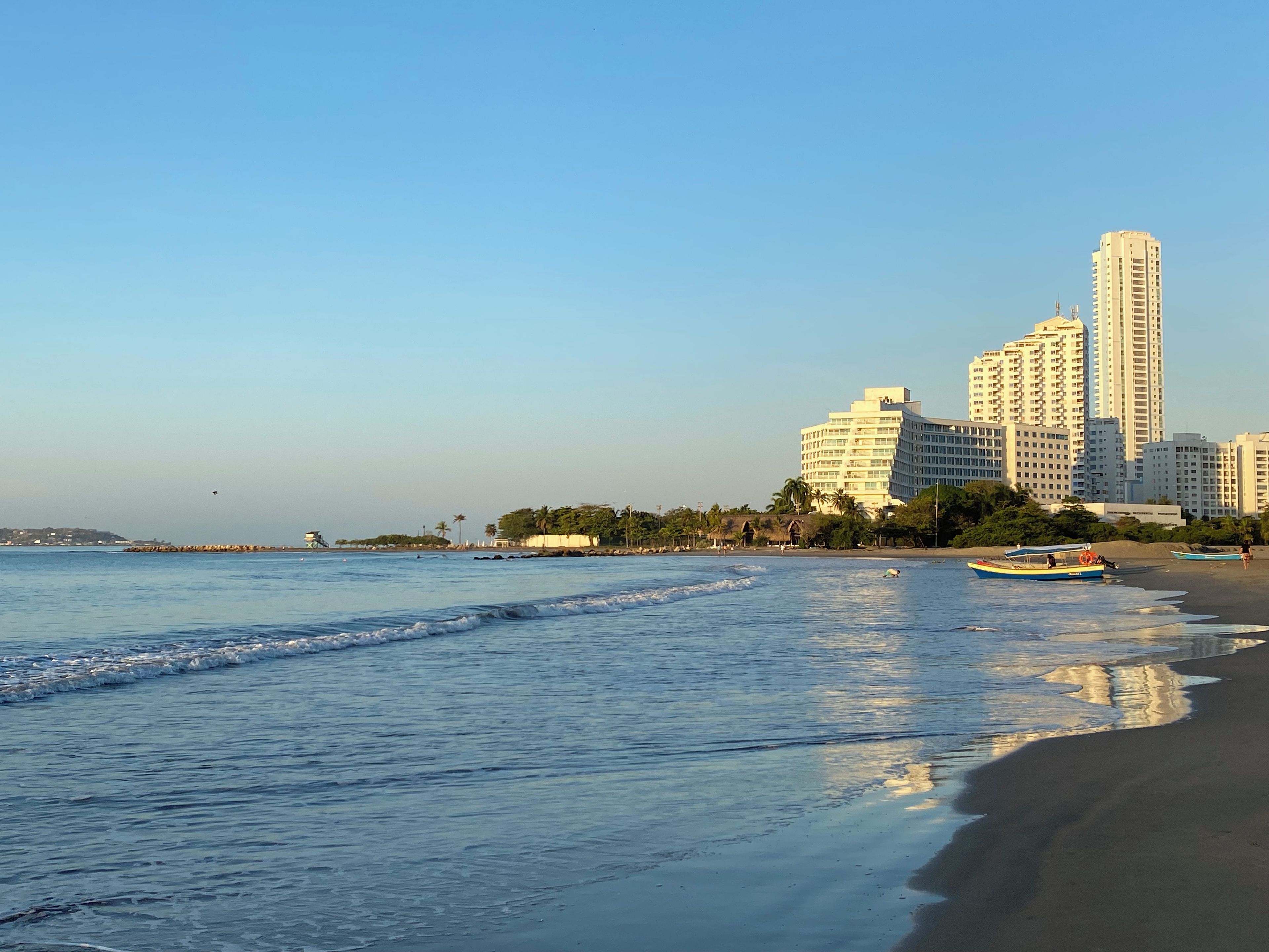A beach with a clear sky, calm sea, and a row of tall buildings in the background.