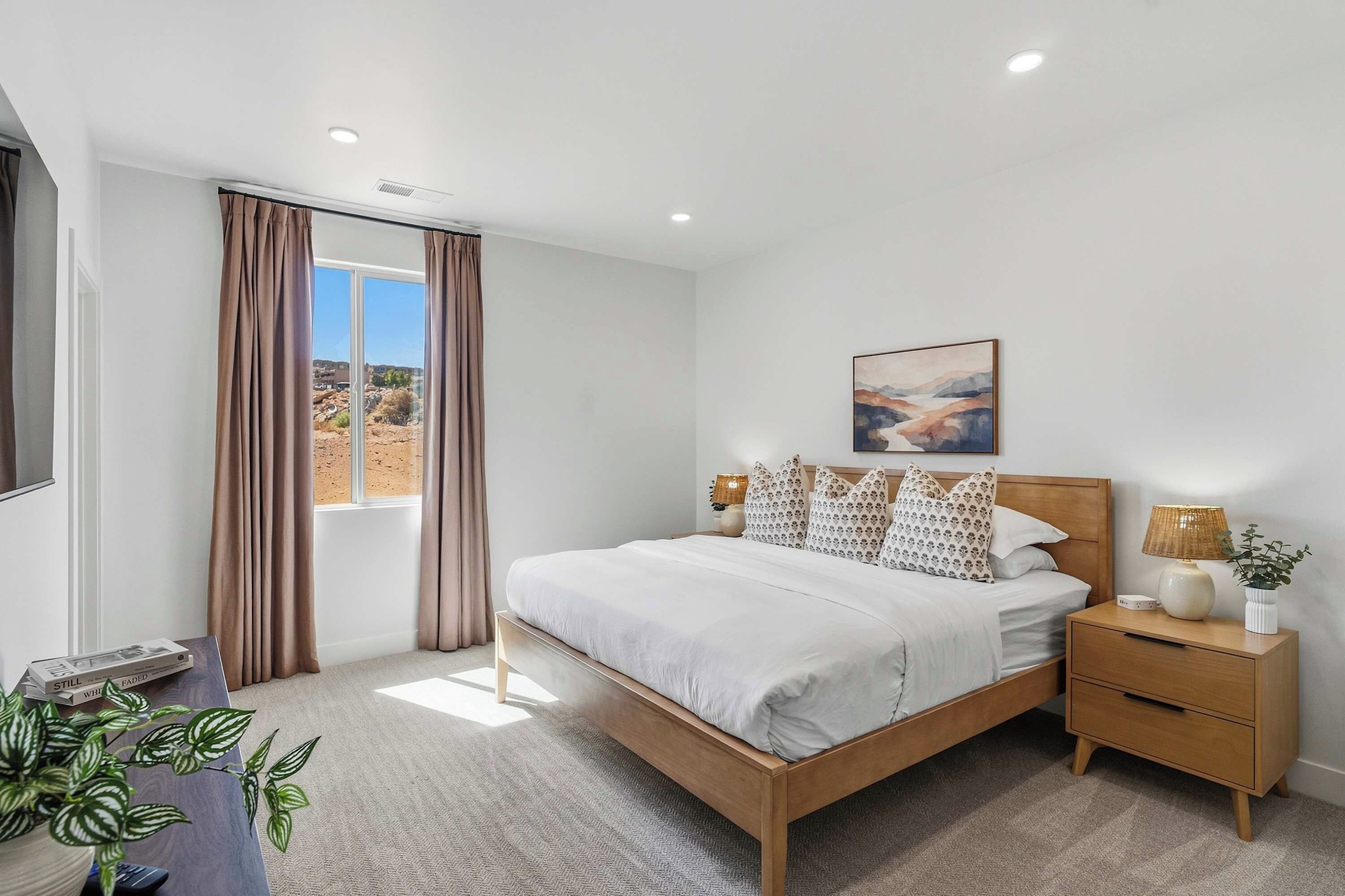 Un dormitorio con cama, mesitas de noche, lámpara, planta, ventana con cortinas y un cuadro en la pared.