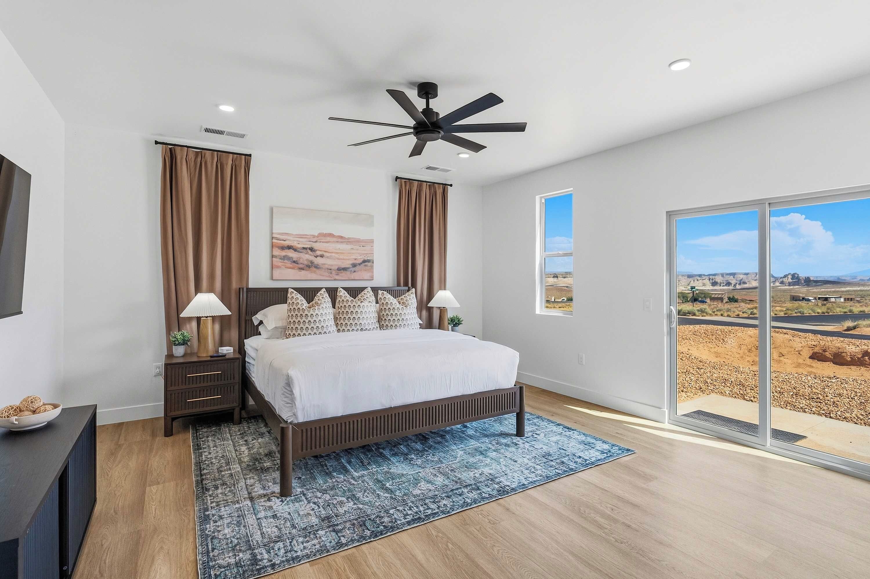 A bedroom with a bed, nightstands, a ceiling fan, a large window with a view, and a painting on the wall.