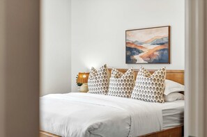 Ein ordentlich bezogenes Bett mit weißer Decke und gemusterten Kissen, ein an der Wand hängendes Landschaftsbild und eine Nachttischlampe.