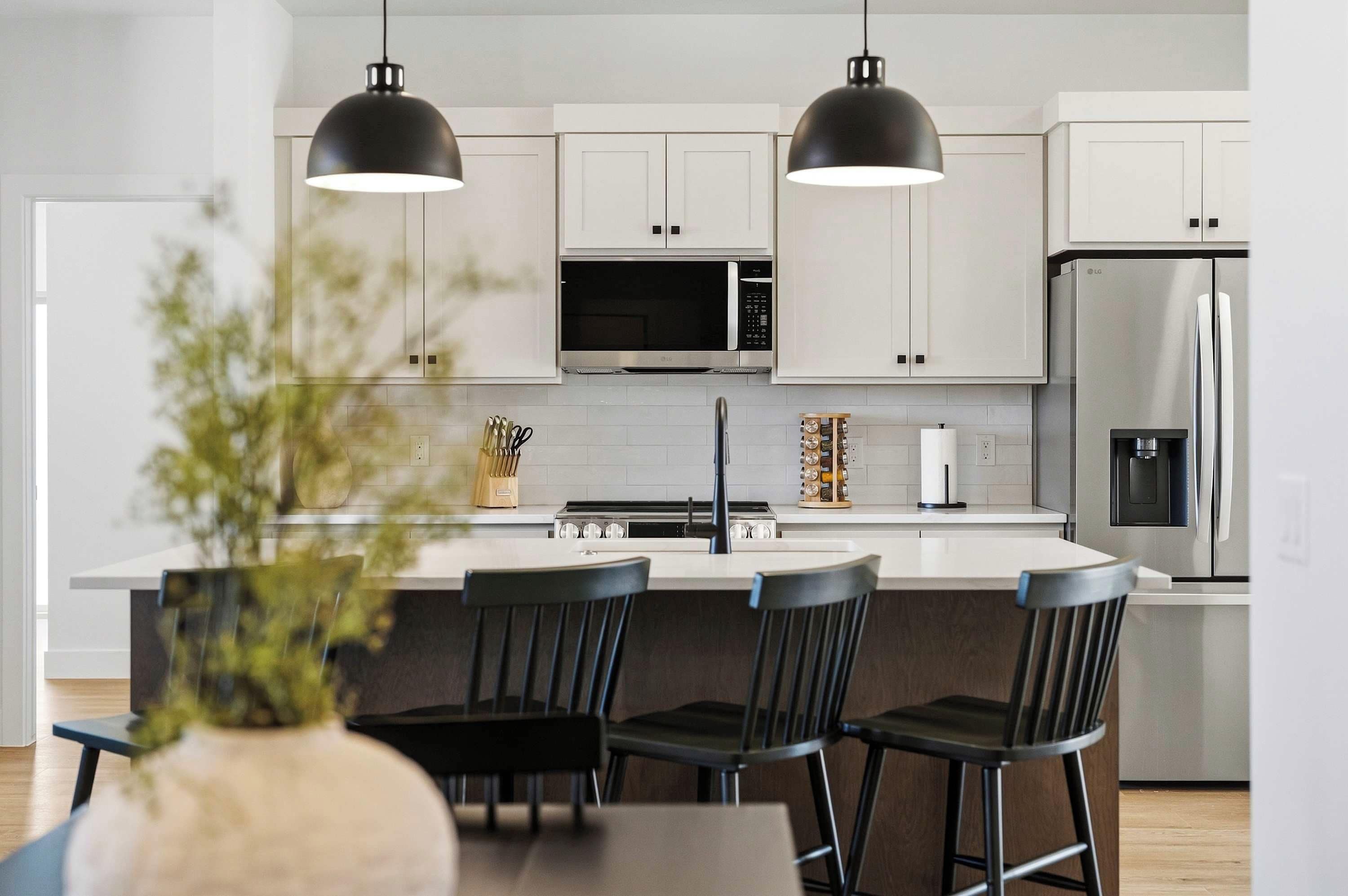 Una cocina moderna con armarios blancos, un refrigerador de acero inoxidable, un microondas y una estufa a gas. Hay tres taburetes negros y una mesa de comedor con un jarrón de flores.