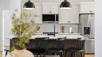 Una cocina moderna con armarios blancos, un refrigerador de acero inoxidable, un microondas y una estufa a gas. Hay tres taburetes negros y una mesa de comedor con un jarrón de flores.