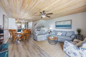 A spacious living room with a wooden ceiling, a dining area with a round table and chairs, a grey sofa set, and a large painting of the ocean. - Coastal Retreat at Beachwalk, Steps from the Gulf (Panama City Beach)