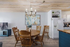 Un coin repas avec une table et des chaises en bois, un lustre et une cuisine dotée de meubles blancs et d’appareils électroménagers.