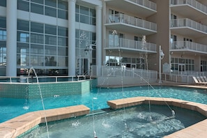 Ein Schwimmbecken mit Wasserspielen, umgeben von einem Gebäude mit mehreren Balkonen und großen Fenstern.