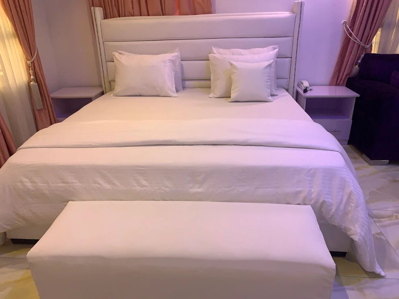Una cama bien tendida con sábanas y almohadones blancos, una mesita de noche con lámpara y un pequeño mueble auxiliar con un teléfono.