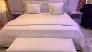 Una cama bien tendida con sábanas y almohadones blancos, una mesita de noche con lámpara y un pequeño mueble auxiliar con un teléfono.
