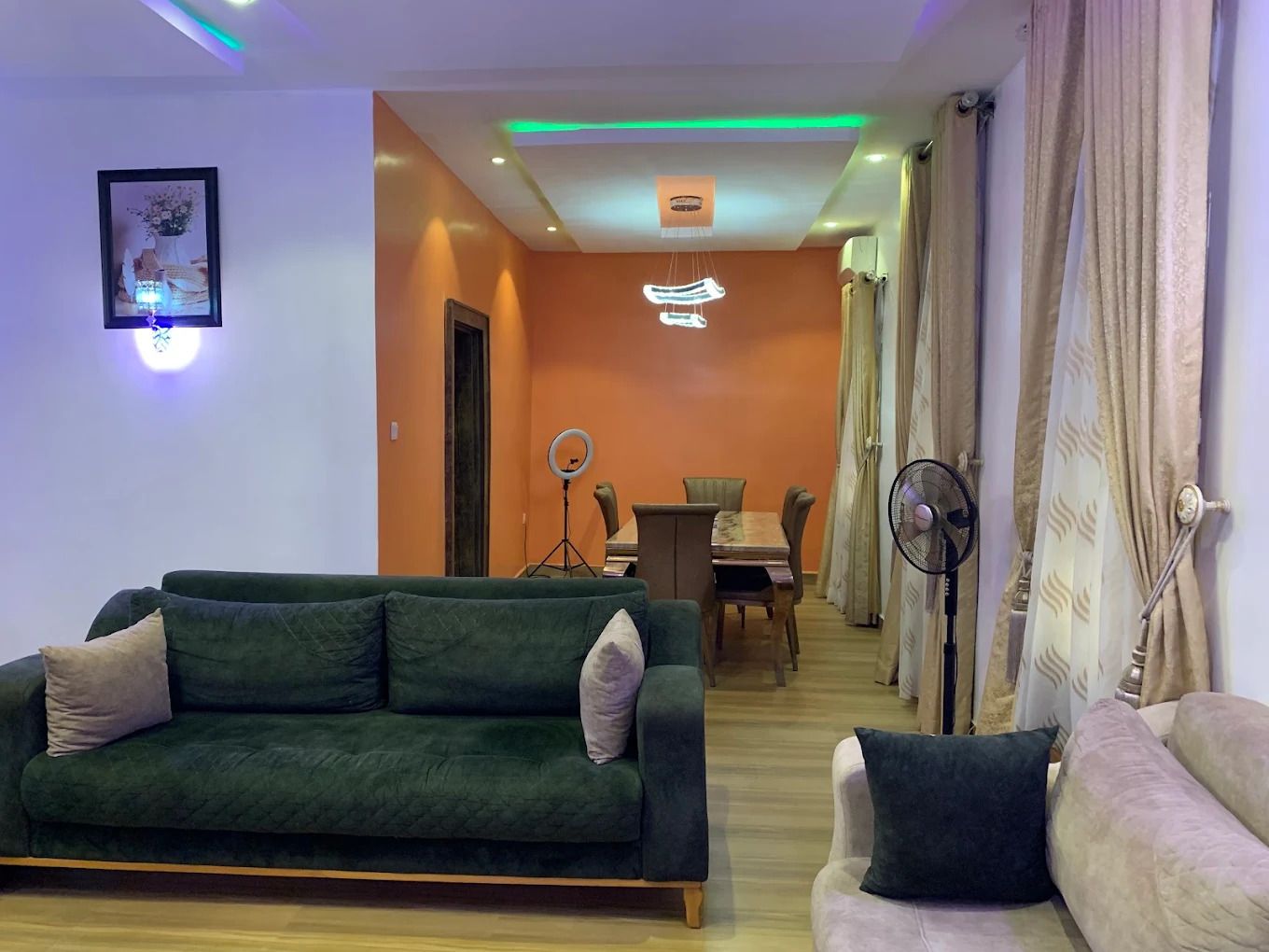 A living room with a green sofa, a white sofa, a dining table with chairs, and a ring light.