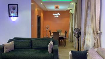 A living room with a green sofa, a white sofa, a dining table with chairs, and a ring light.