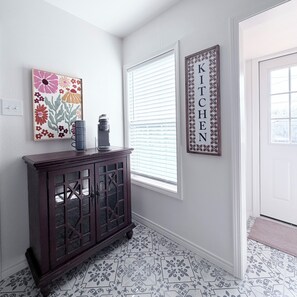 A hallway with a dark wooden cabinet, a framed floral artwork, a framed word 'KITCHEN' on the wall, and patterned floor tiles. - Beautiful, 2-bedroom house with AC, fast WiFi, quiet street, close to downtown. (Corpus Christi)