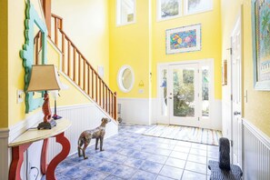 A bright yellow hallway with a wooden staircase, a round mirror, and a dog standing on the floor. - Soundfront w/ Hot Tub, Game Room & Resort Perks! (Corolla)