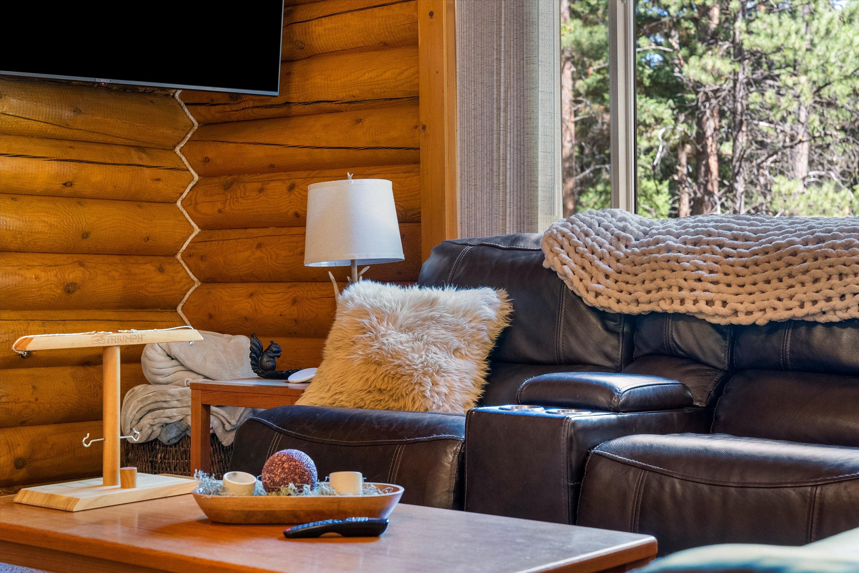 A cozy living room with a leather sofa, a knitted throw blanket, a wooden coffee table with a bowl of snacks, a lamp, and a television mounted on a wooden wall.
