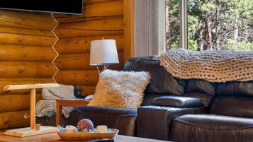 A cozy living room with a leather sofa, a knitted throw blanket, a wooden coffee table with a bowl of snacks, a lamp, and a television mounted on a wooden wall.