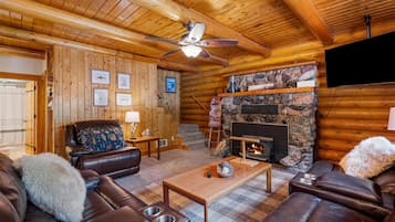 A log cabin interior with a fireplace, leather furniture, wooden walls, and a ceiling fan.