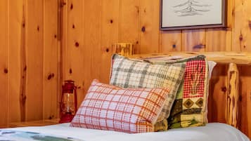 A wooden cabin room with a bed, pillows, a lantern, and a framed drawing on the wall.