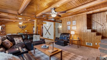 A log cabin interior with a leather sofa, a wooden coffee table, a ceiling fan, and a staircase.
