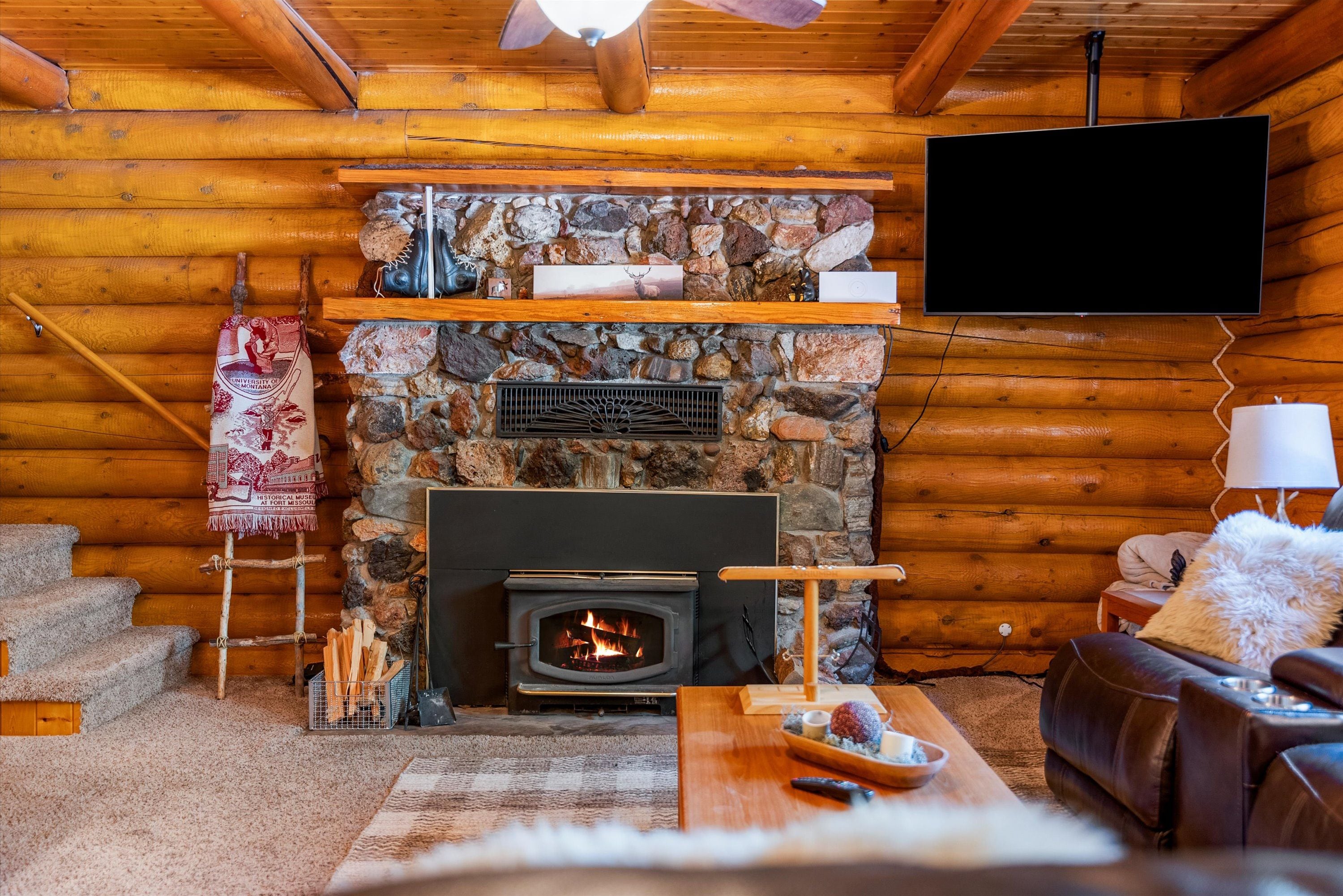 A log cabin interior with a stone fireplace, wooden walls, a TV mounted above the fireplace, a sofa, and a dining table with a tray.