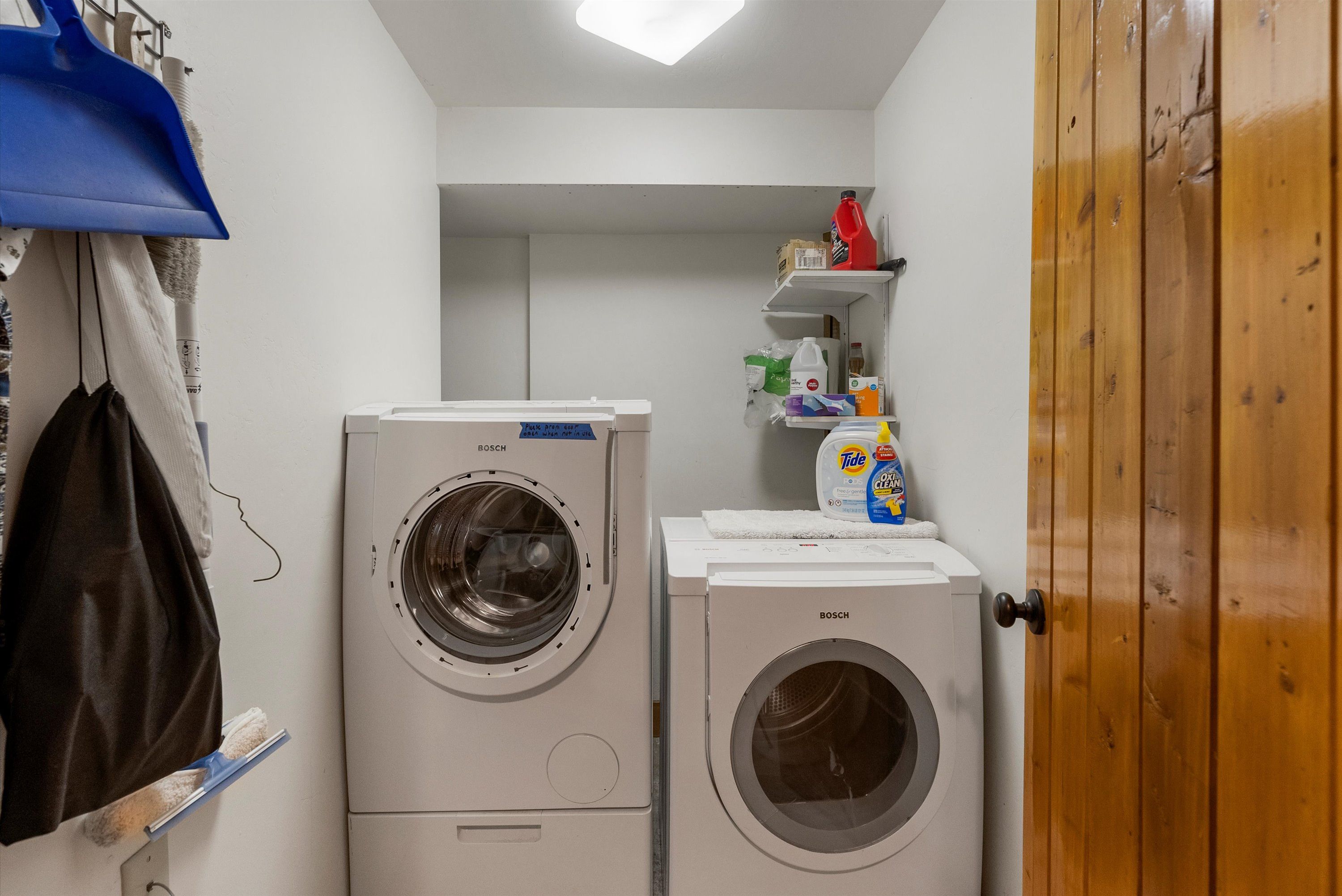 A laundry room with two Bosch washing machines, a shelf with cleaning supplies, and a wooden door.