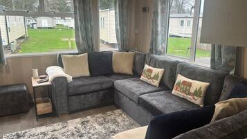 A modern living room with a sectional sofa, a small table, and a view of a grassy area with mobile homes.