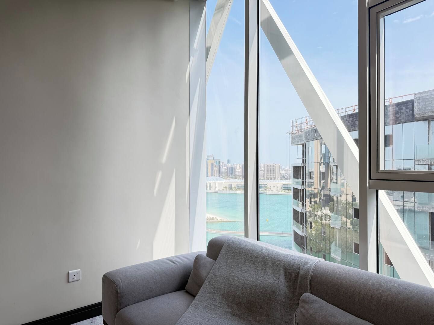 A modern living room with a grey sofa, a large window offering a view of a cityscape and water body.