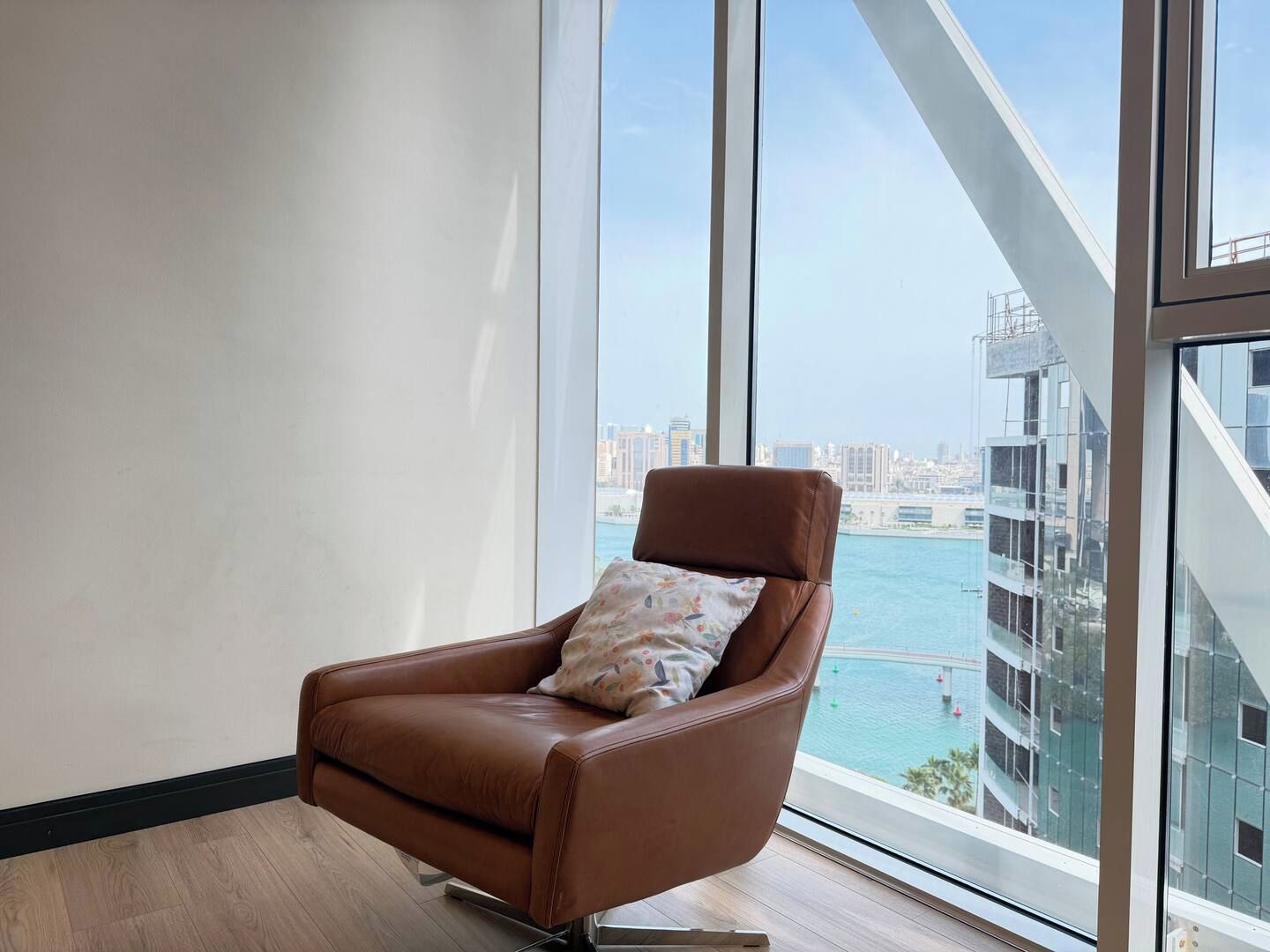 A brown leather armchair with a floral pillow in a room with large windows overlooking a cityscape and water.