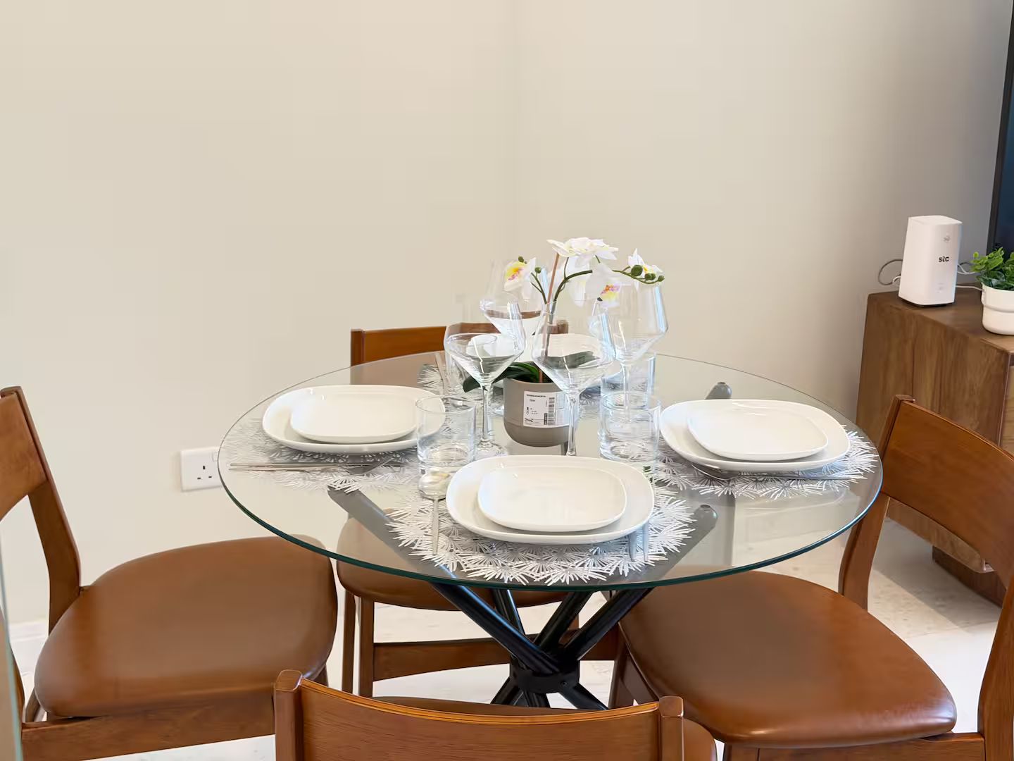 Une table de salle à manger ronde en verre, avec de l’assiette blanche, de la verrerie transparente et un vase contenant des fleurs blanches.