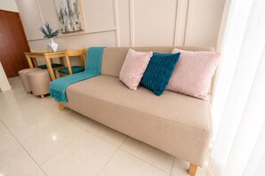 Ein beiges Sofa mit Türkis- und Rosakissen, ein kleiner Holztisch mit einer Vase und eine türkisfarbene Decke.