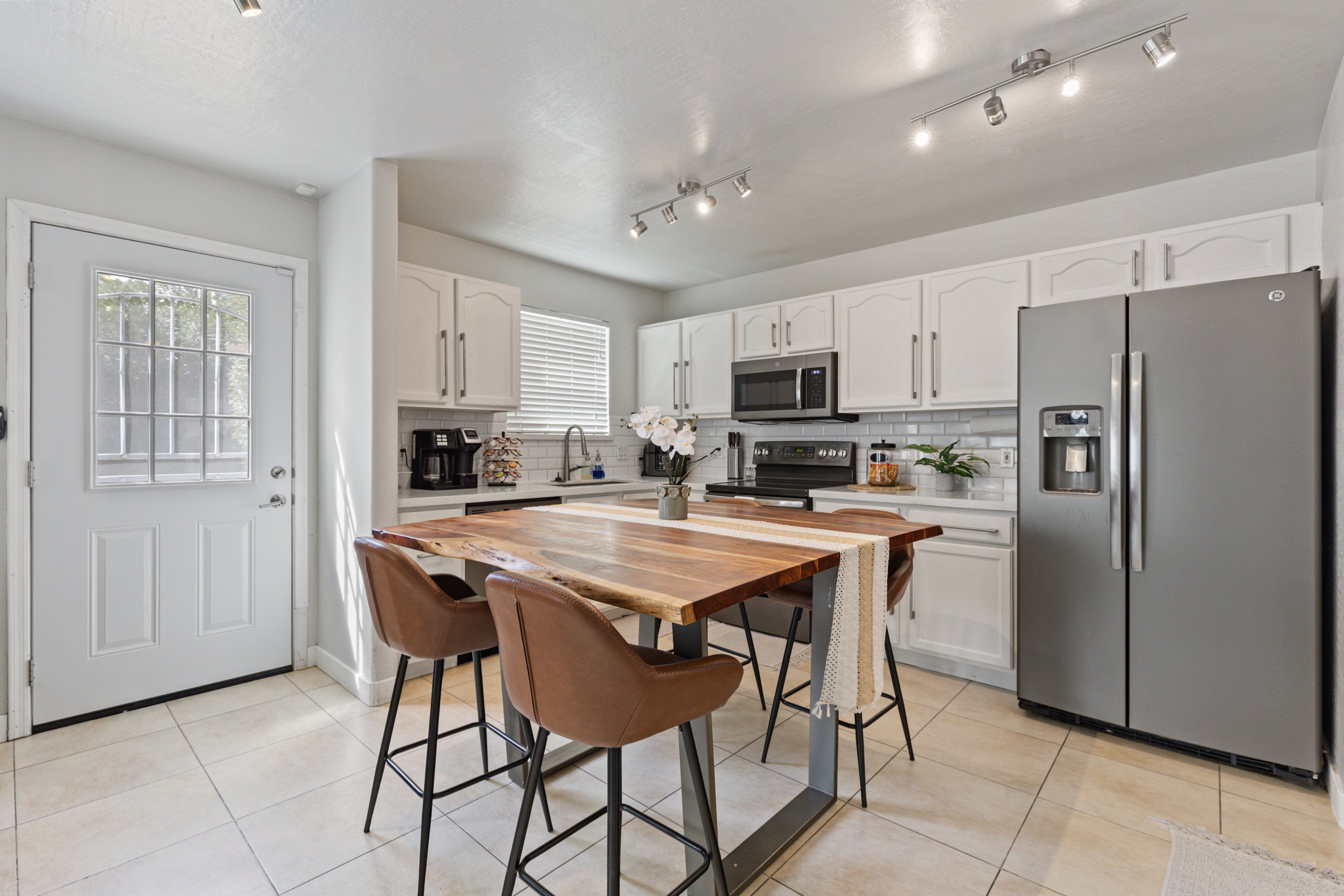 A modern kitchen with white cabinetry, a stainless steel refrigerator, a wooden island with bar stools, and a window with blinds.