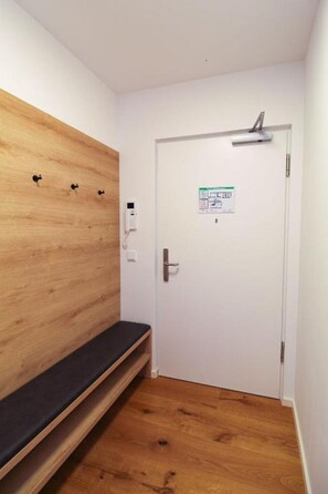 Ein Zimmer mit einer hölzernen Sitzbank, einer weißen Tür mit einem Aushang und einem kleinen Regal.