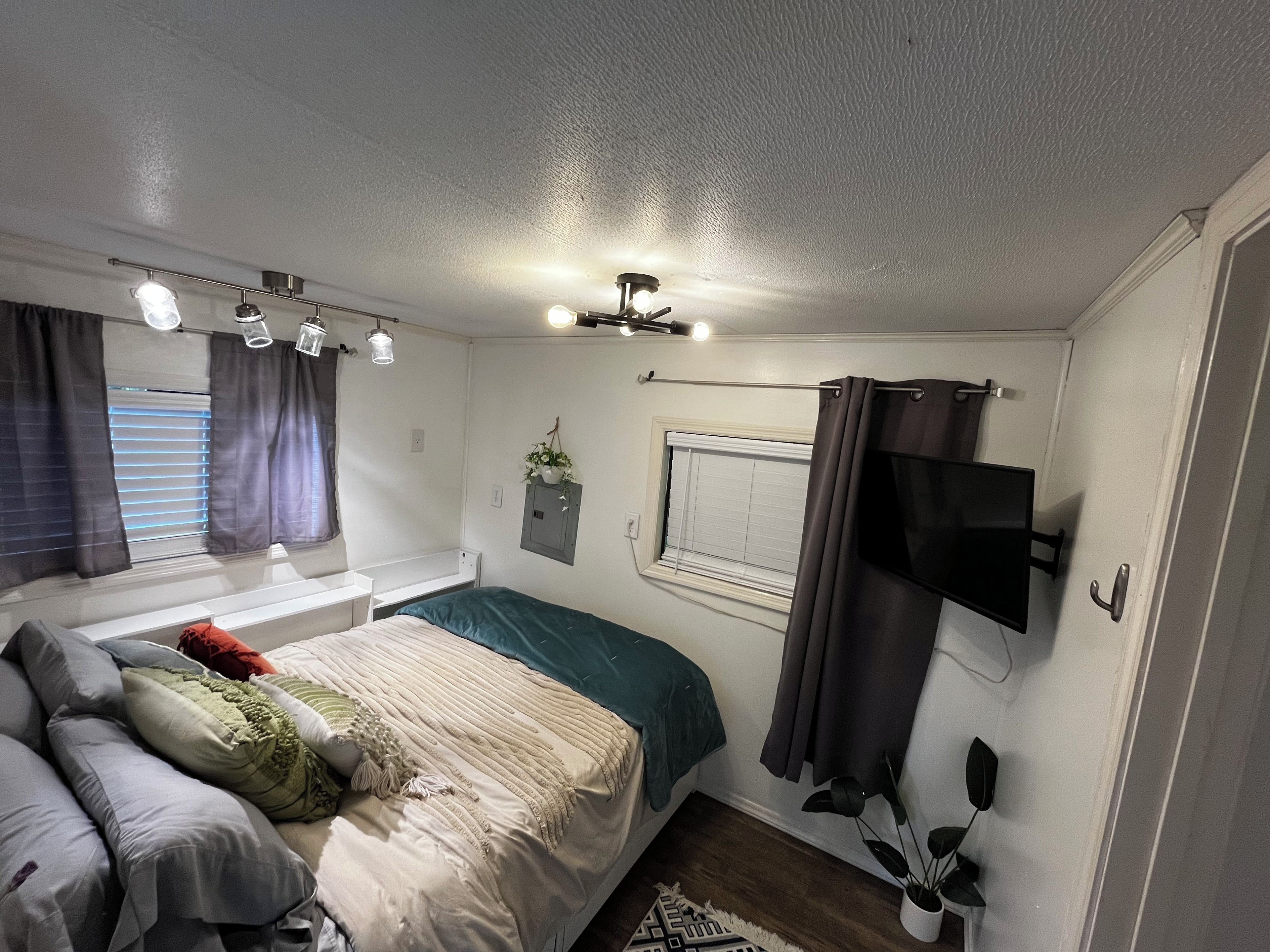 A bedroom with a bed, a window with curtains, a ceiling fan, and a mounted television.