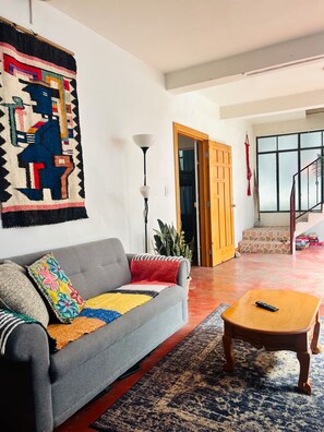 A living room with a grey sofa, colorful cushions, a wooden coffee table, a patterned rug, a wall hanging, a floor lamp, and a staircase leading to an upper level. - Casa Bentley: 360° rooftop terrace + lush garden (San Pedro La Laguna)