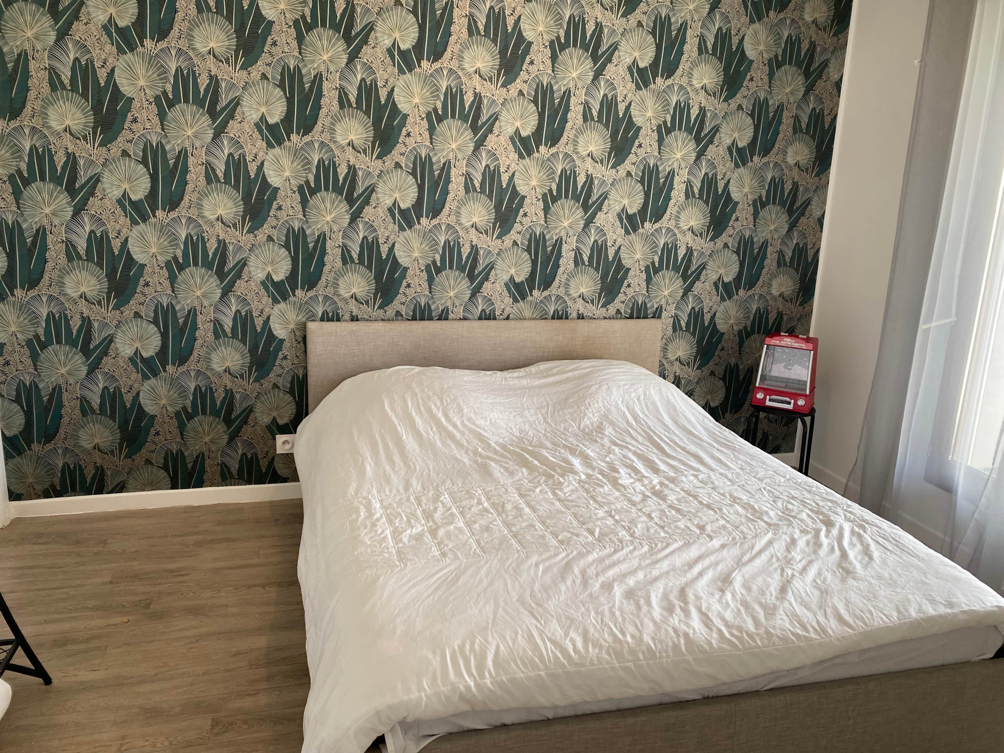 Een bed met een witte spreideken in een kamer met behang met een patroon en een klein rood voorwerp op een voetstuk.