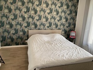 Een bed met een witte spreideken in een kamer met behang met een patroon en een klein rood voorwerp op een voetstuk.
