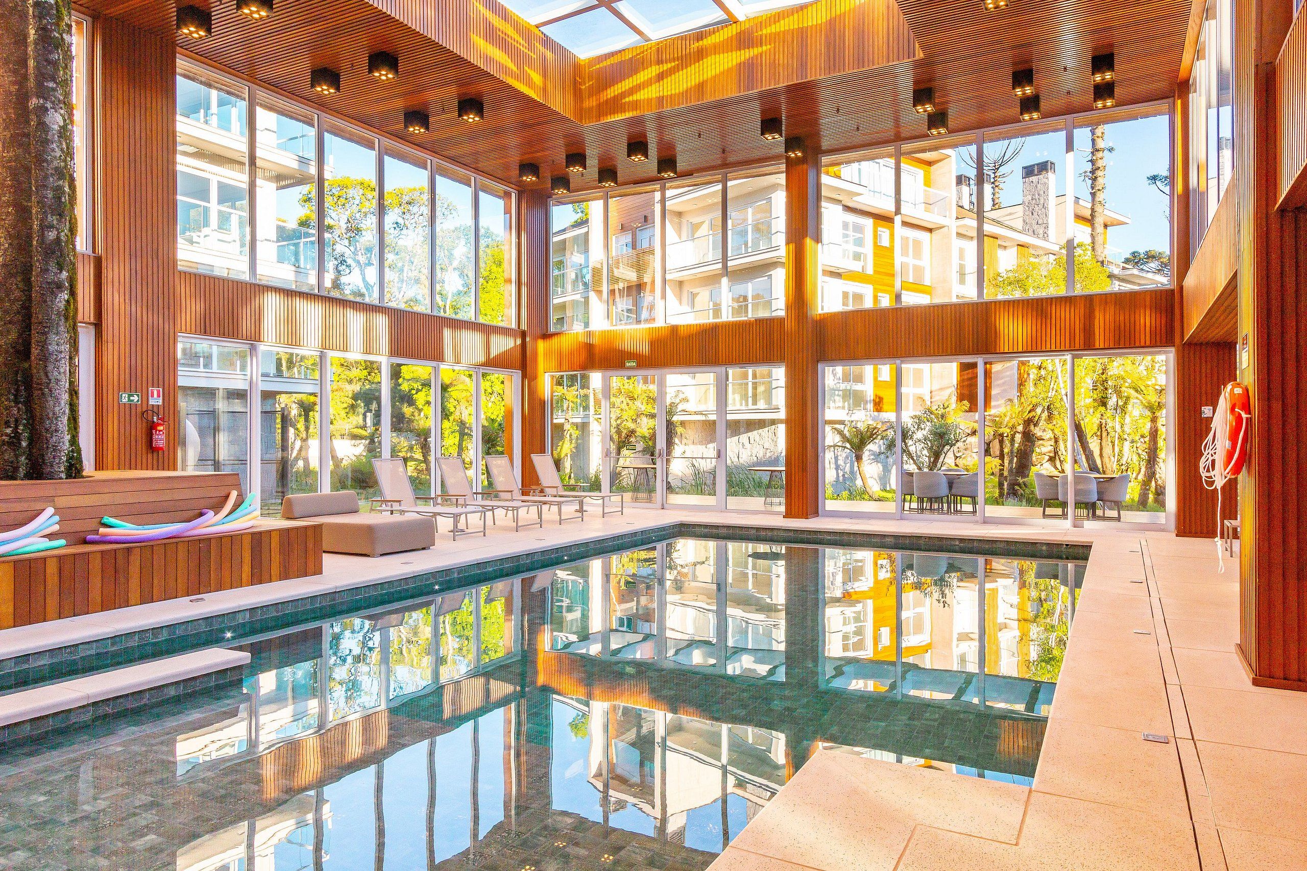 A modern indoor pool area with clear glass walls, wooden paneling, and lounge chairs.