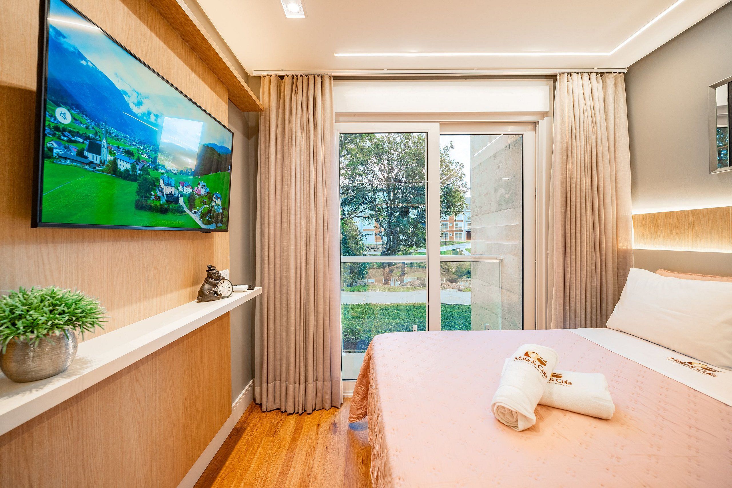 A modern hotel room with a large TV, a bed with white linens, a small plant, and a view of a residential area through the window.
