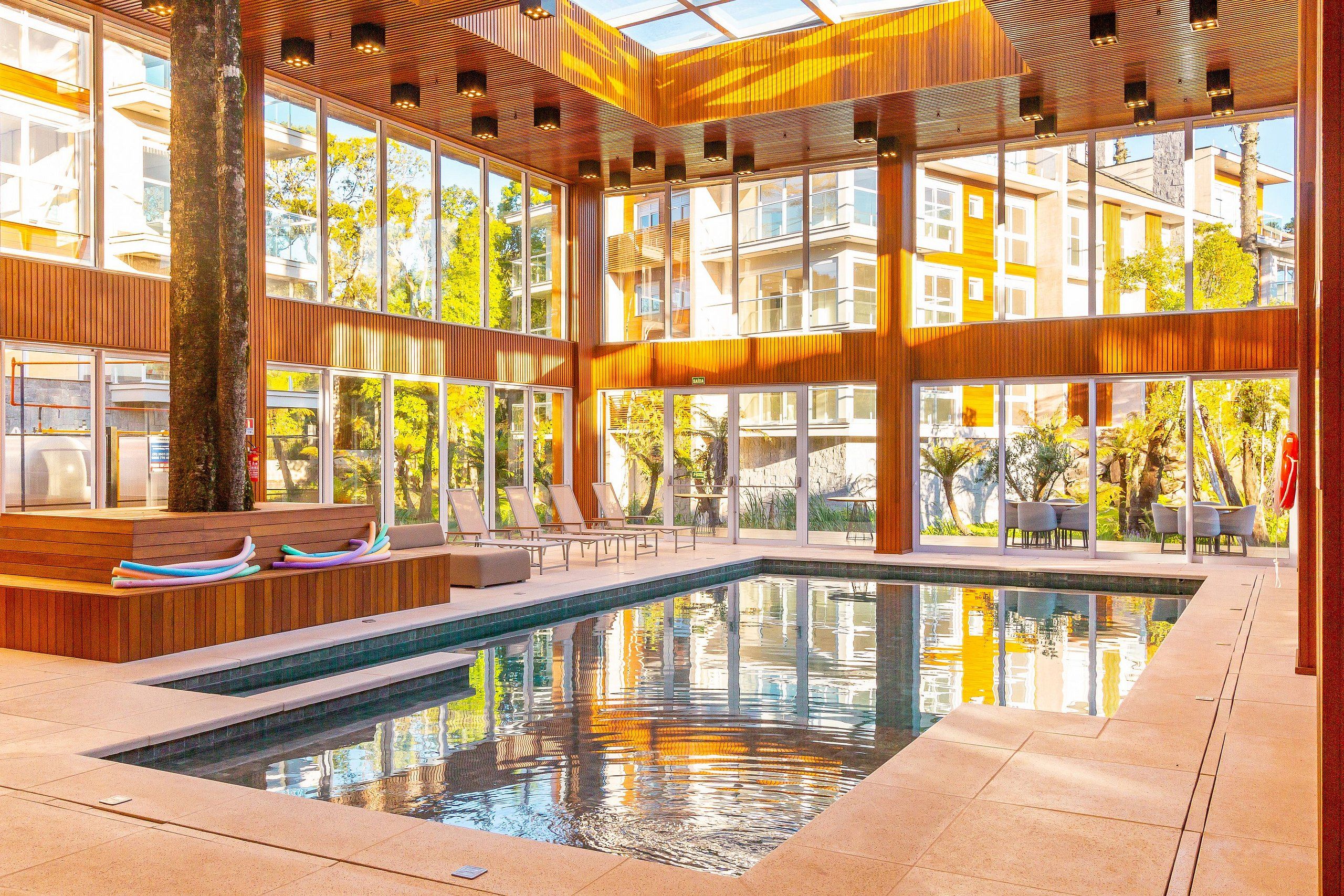 Un área de piscina interior moderna con techo de vidrio, paneles de madera y asientos junto a la piscina.