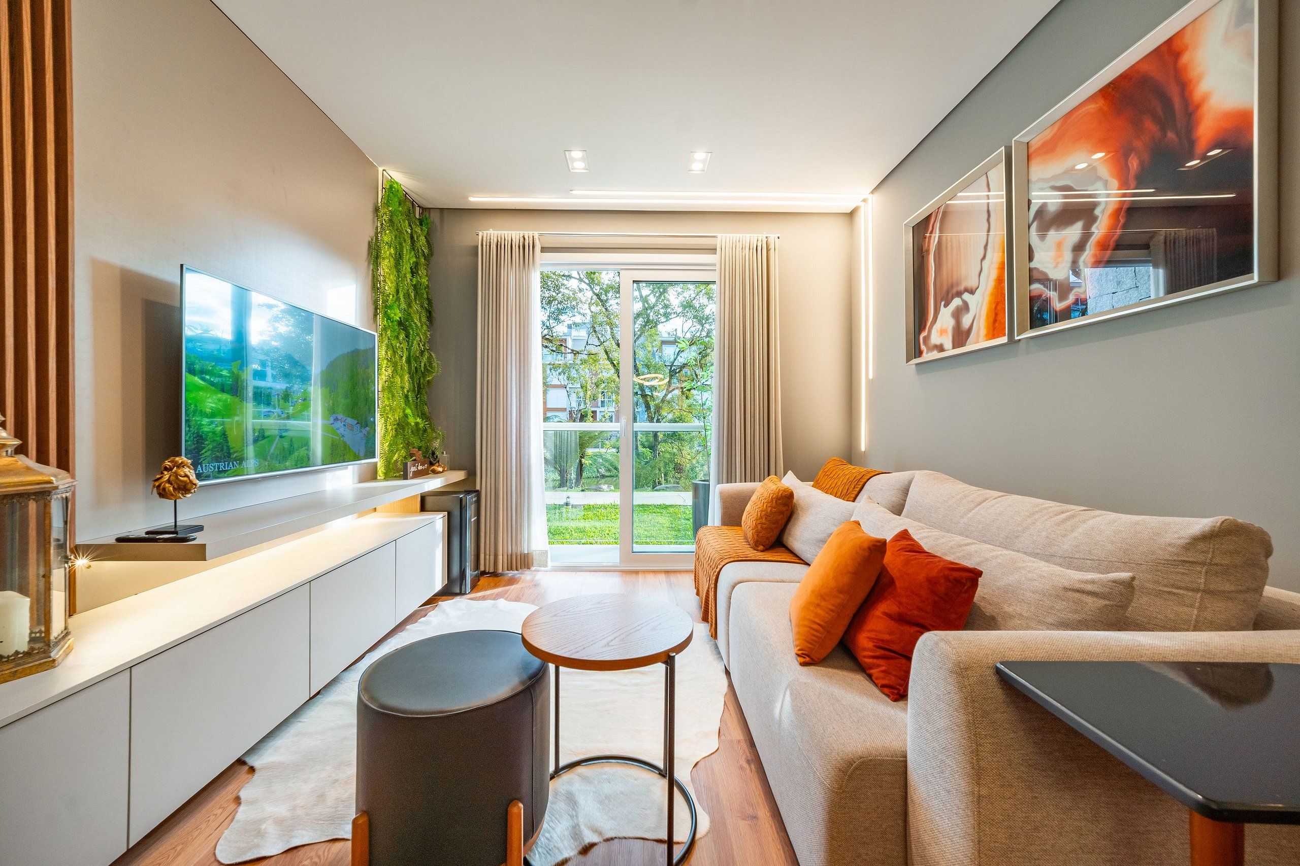 Ein modernes Wohnzimmer mit einem Flachbildfernseher, einer Couch mit Kissen, einem kleinen runden Tisch und einem großen Fenster mit Vorhängen.