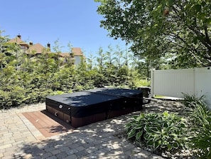 A hot tub in a garden with a paved area, surrounded by greenery and a white fence.