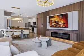 A modern living room with a flat-screen TV mounted on a wall, a fireplace, and a dining area with white chairs and a kitchen in the background. - 7BR house with private pool in clermont (Clermont)